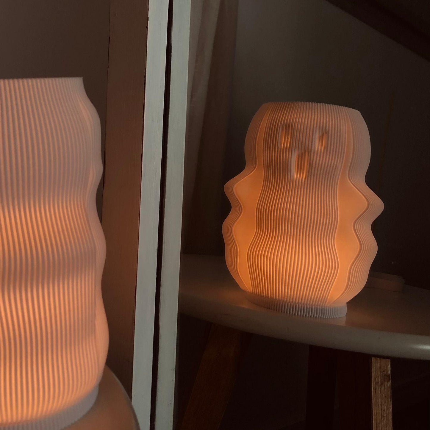 A glowing ghost-shaped lamp sits on a small table, casting a soft, warm light. The lamp’s wavy, ribbed design adds texture and creates a playful, eerie effect as it reflects off a nearby mirror.